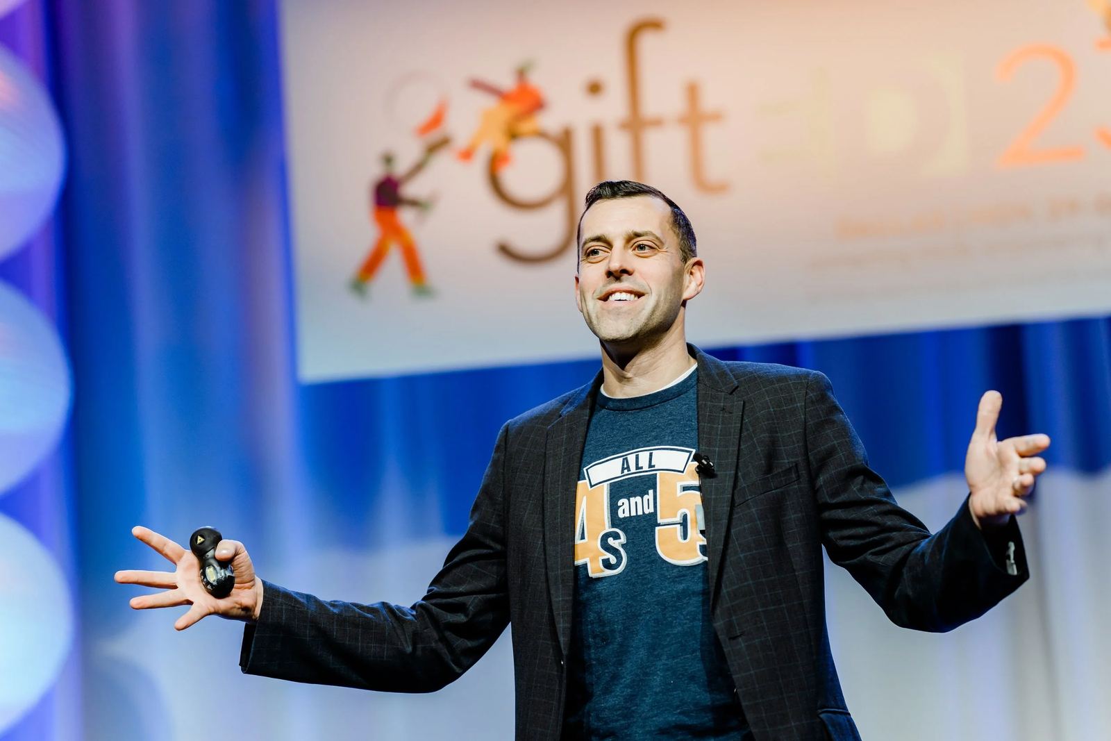 Andrew Sharos on stage, smiling with his arms open as he speaks to a conference audience