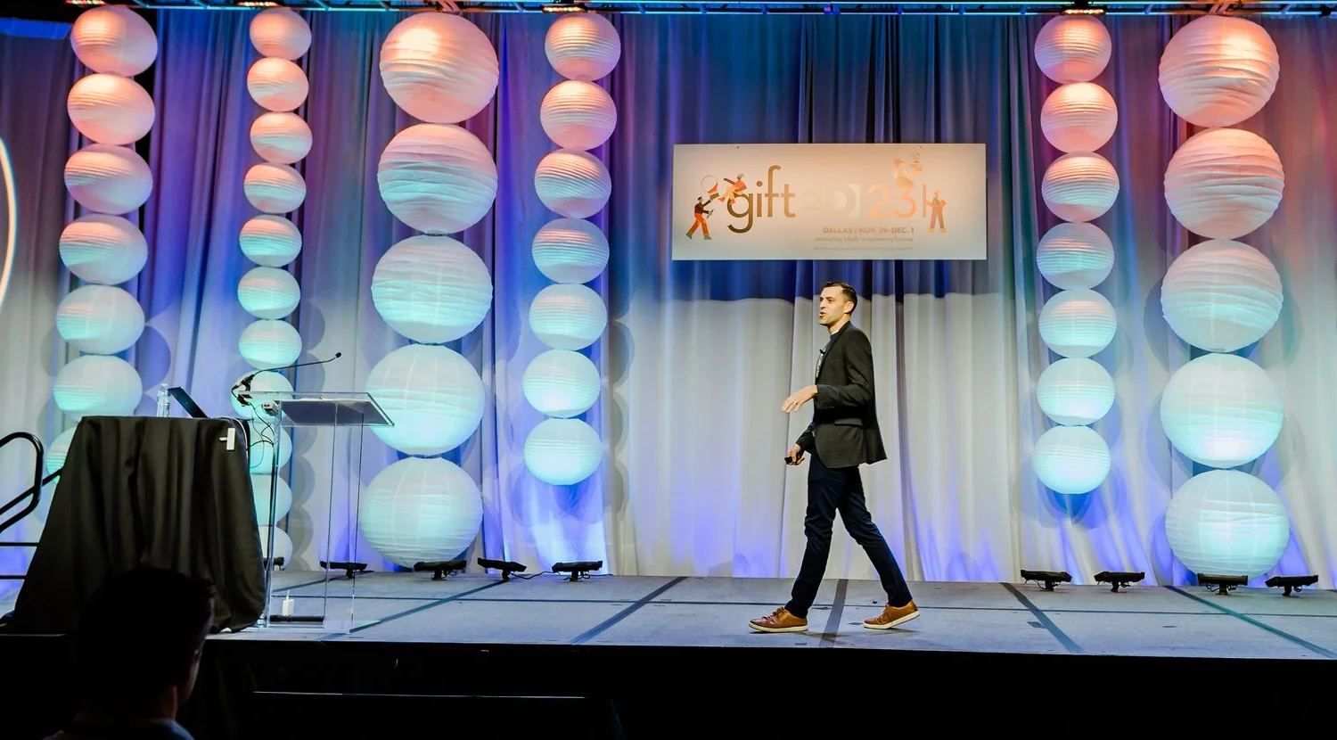Andrew Sharos crossing the stage mid-keynote, in front of a backdrop of lit paper lanterns