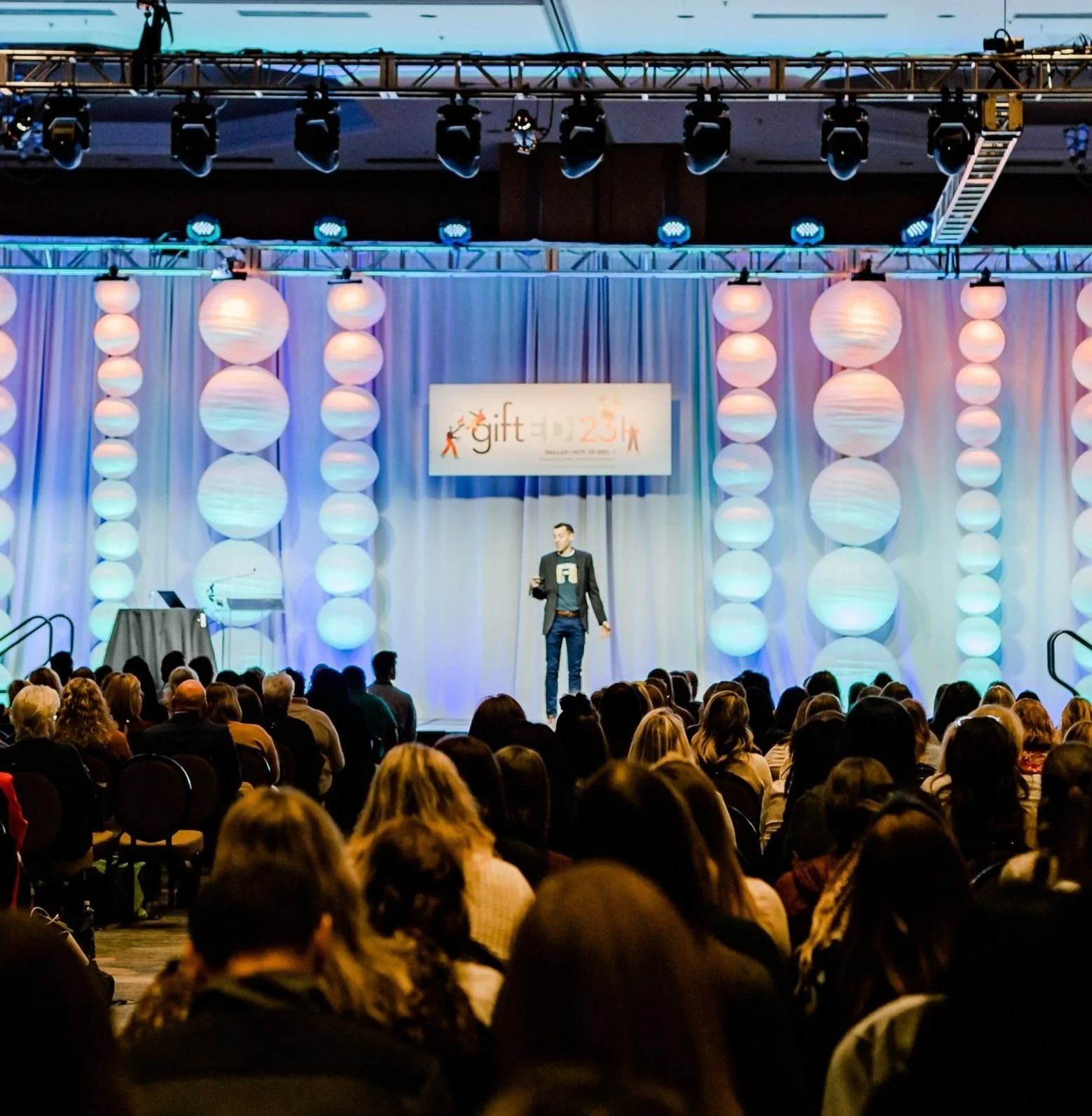 A packed conference hall watching Andrew Sharos speak on a brightly lit stage